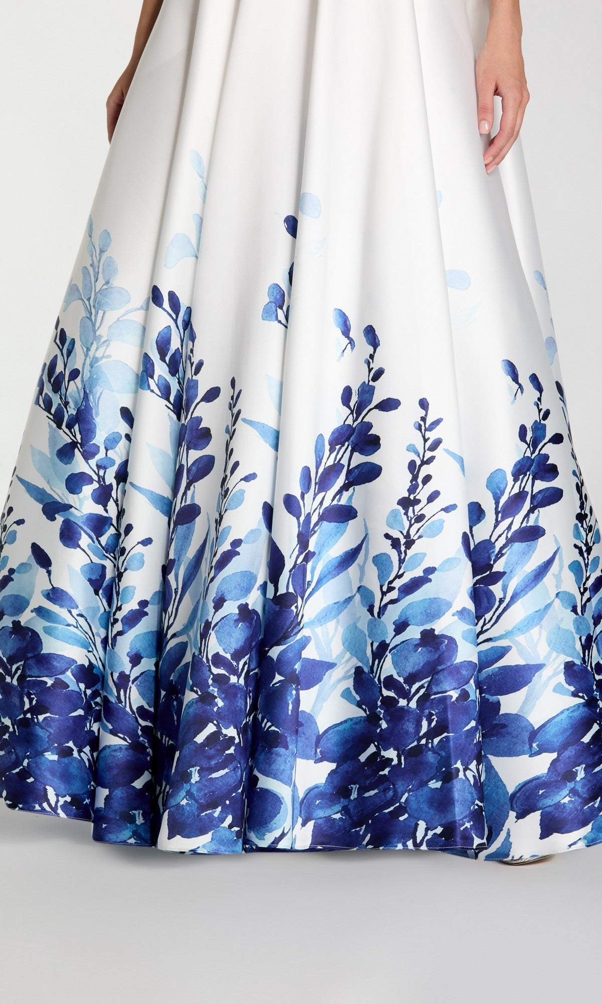 A close-up image shows the lower part of the elegant Mac Duggal 56316 Long Formal Dress—a strapless white Mikado ballgown featuring a blue and navy floral pattern near the hem—and one hand.
